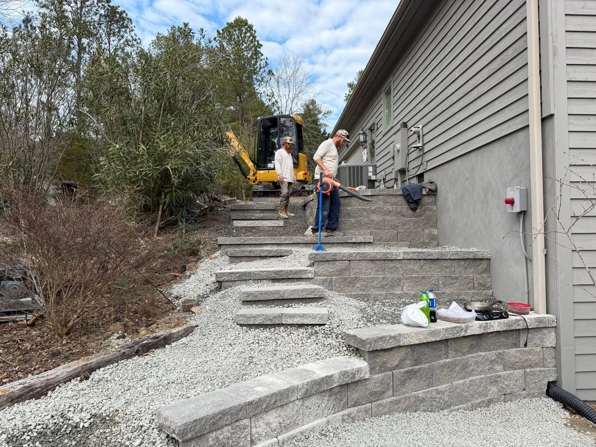 Workers building outdoor concrete stairs beside a house, with a backhoe and tools on site.