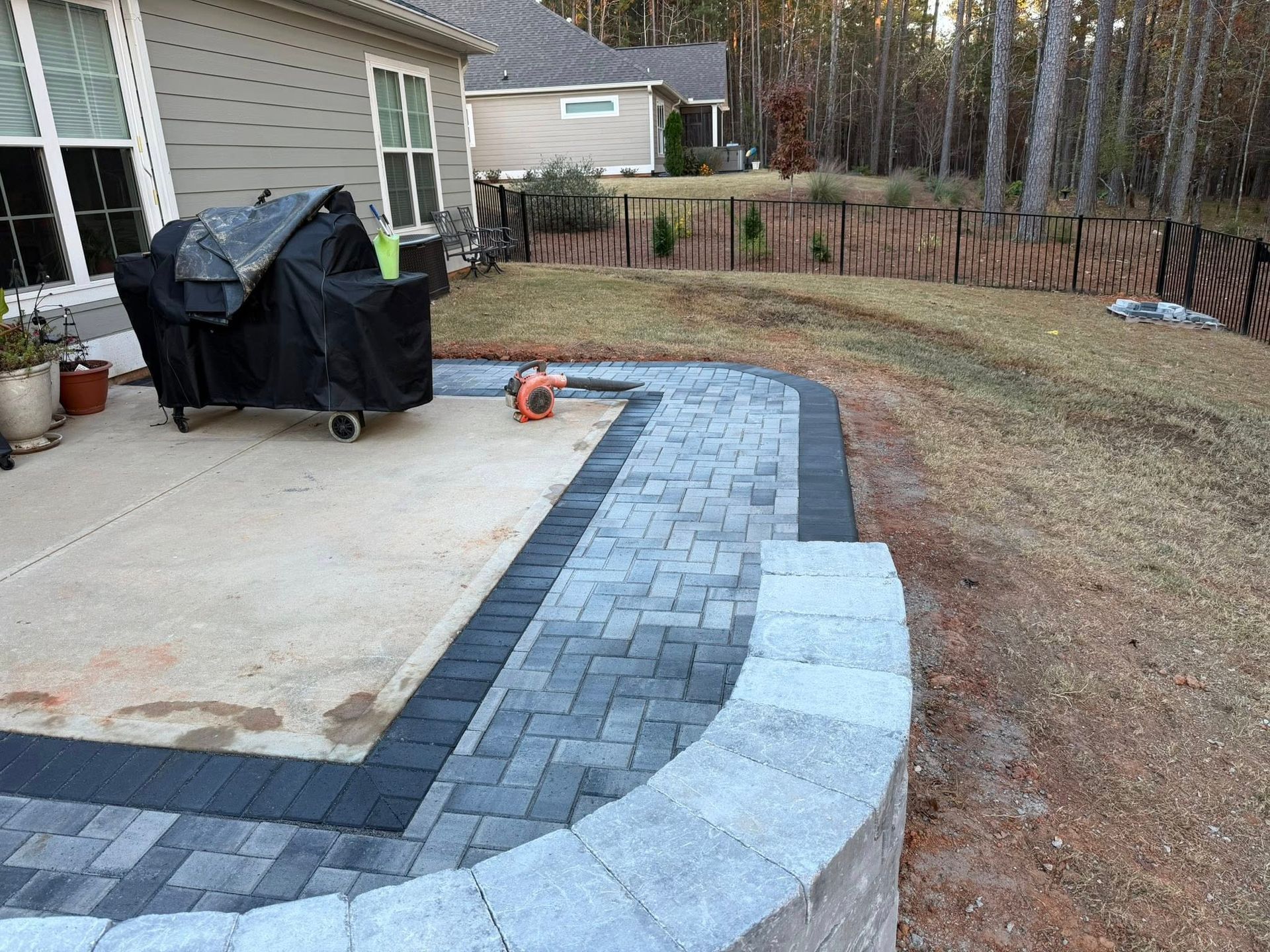 Backyard patio with curved stone paver path, grill, and brown lawn beside a house and woods