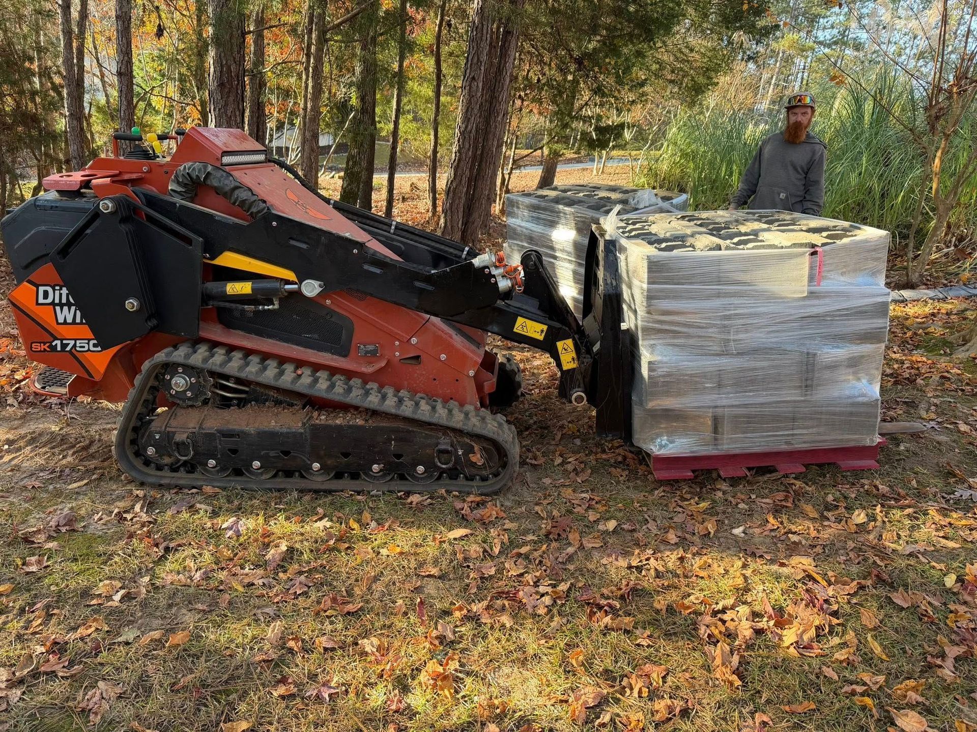 Red and black skid-steer loader lifting a wrapped pallet of blocks in a wooded yard.