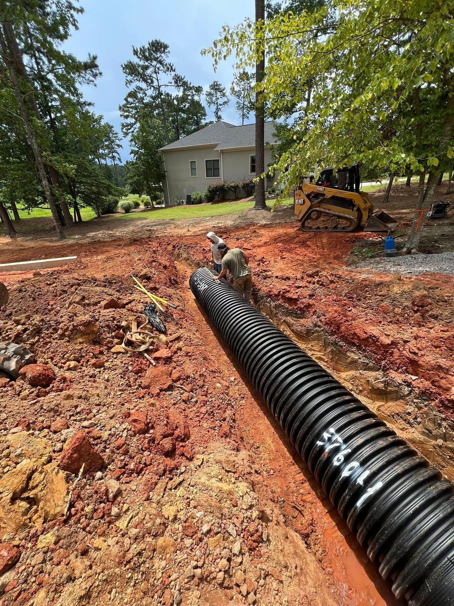 Black corrugated drain pipe laid in a red dirt trench at a rural construction site with a house in the background