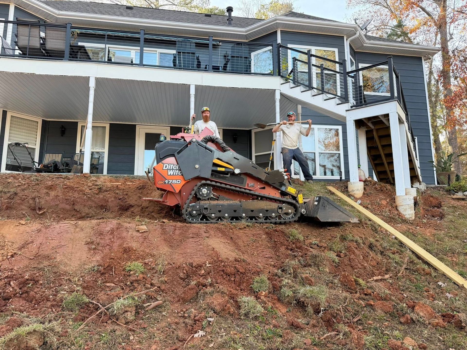 Worker on a tracked machine clearing mud from a steep yard beside a house damaged by a landslide