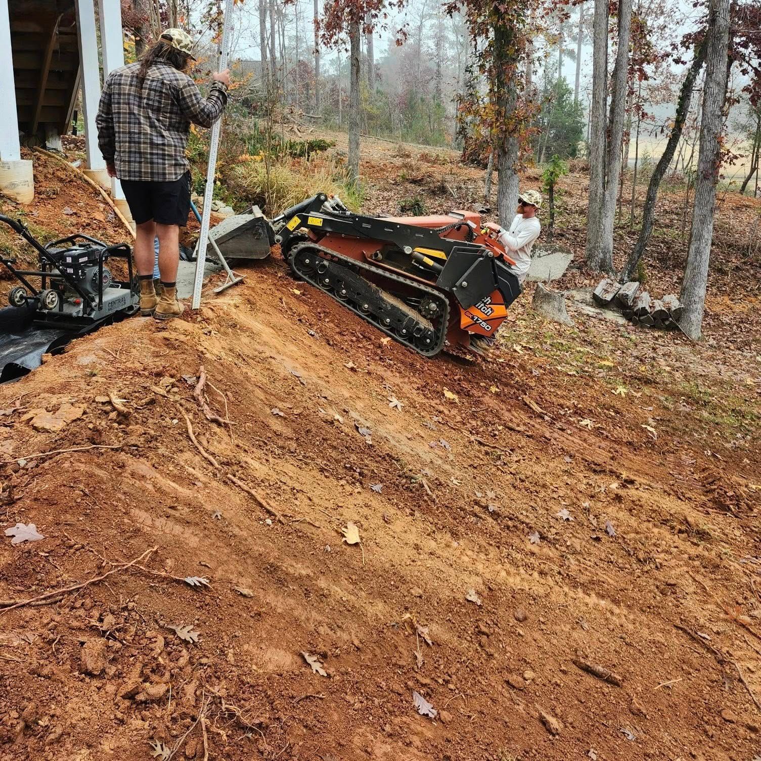Two people grading a dirt trail with a tracked skid steer in a wooded area