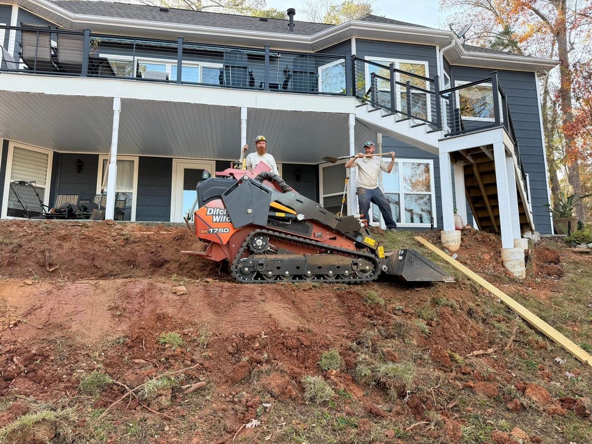 Small excavator on a steep muddy slope beside a house with visible structural damage