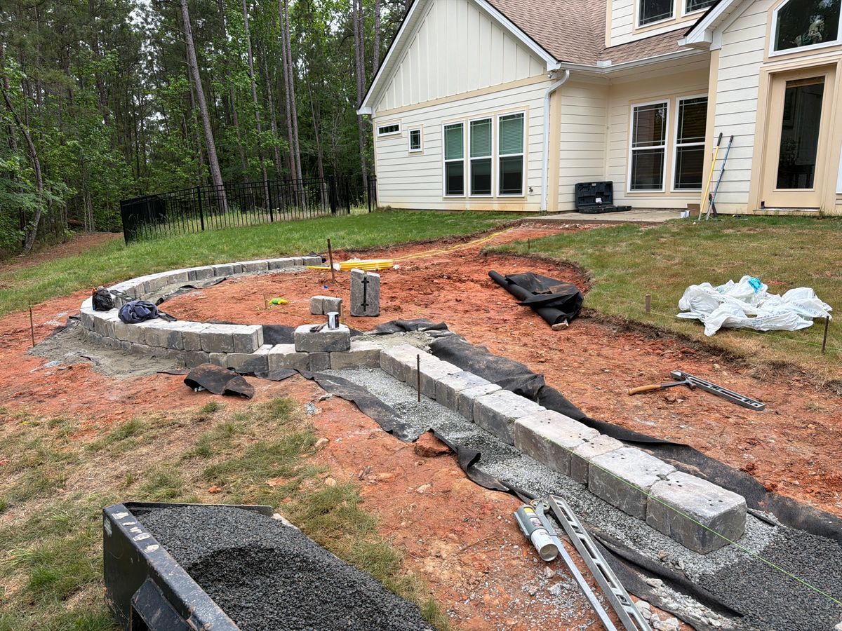Backyard construction with curved concrete retaining wall and dirt trench beside a house and trees