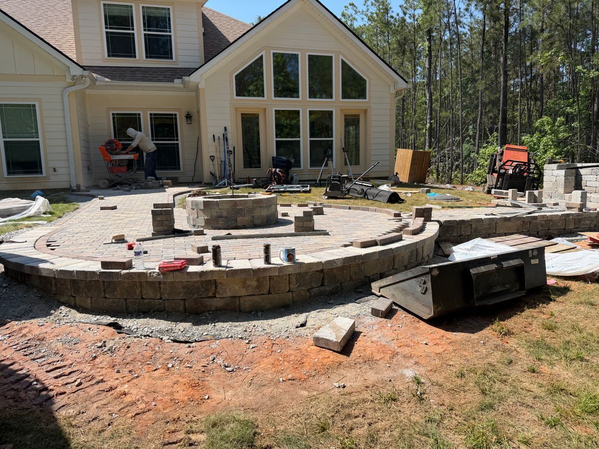 Backyard patio under construction beside a house, with a circular stone fire pit and scattered materials.
