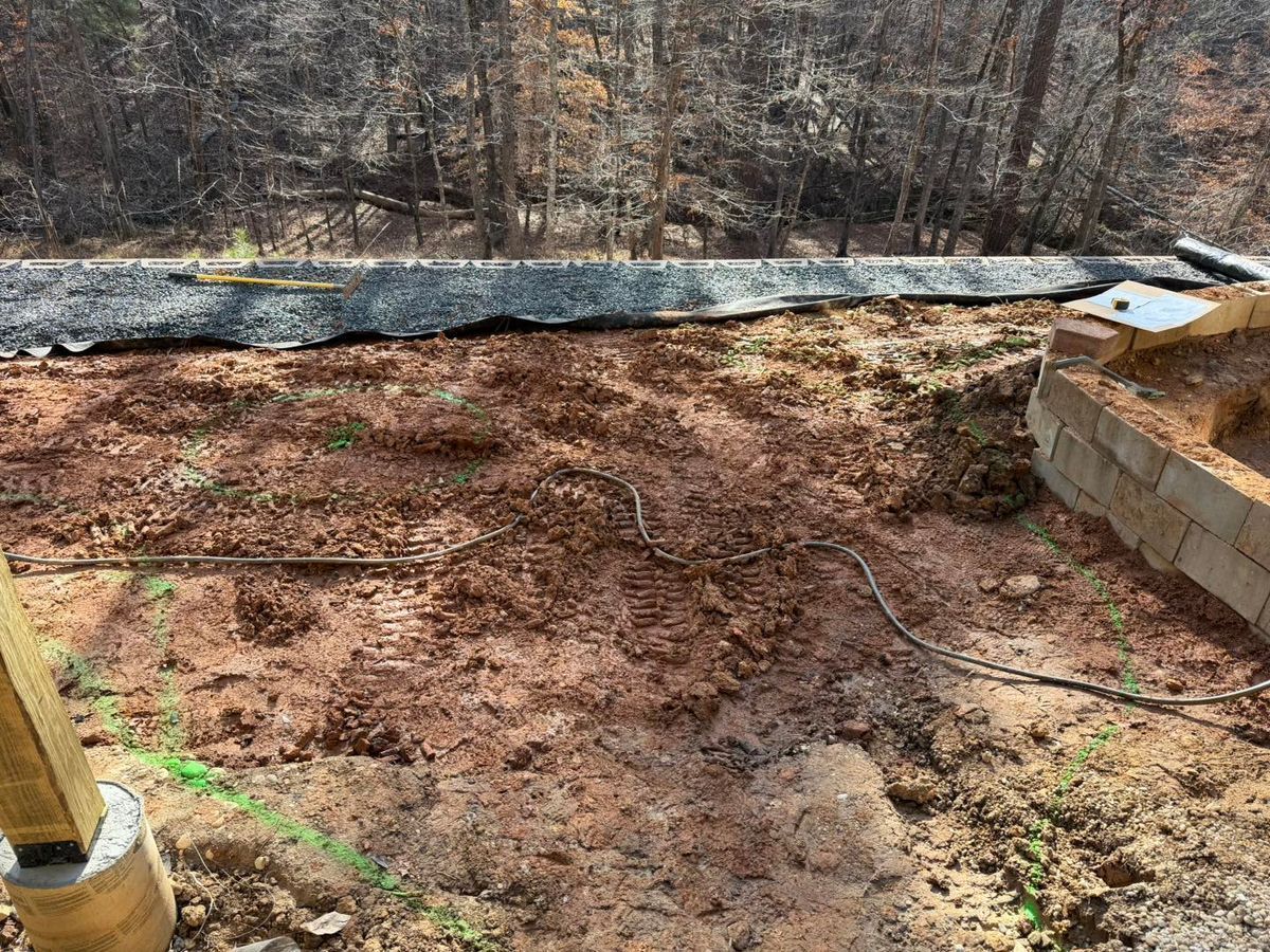 Freshly cleared dirt area beside a retaining wall and wooded slope, with a black barrier at the top