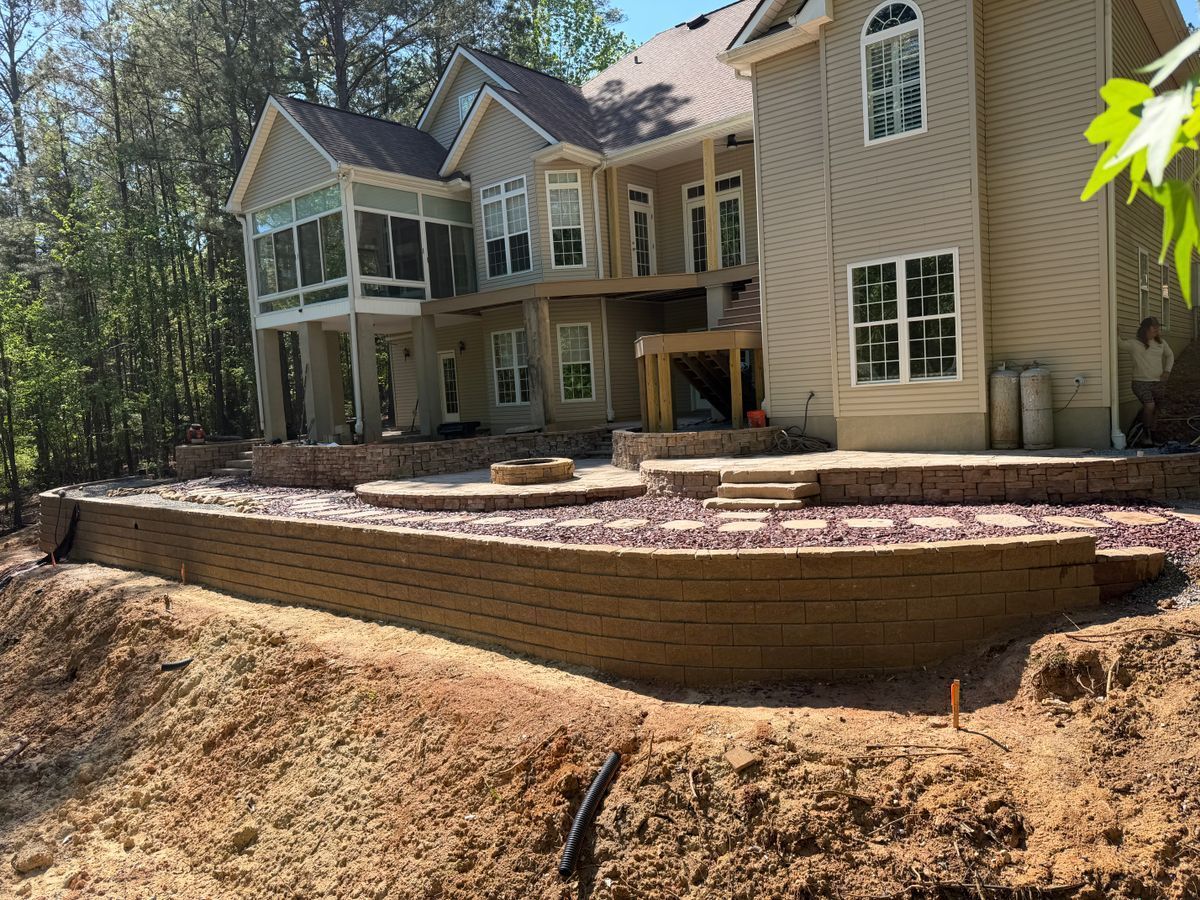 Large beige house with a raised stone patio and retaining wall in a wooded backyard under construction