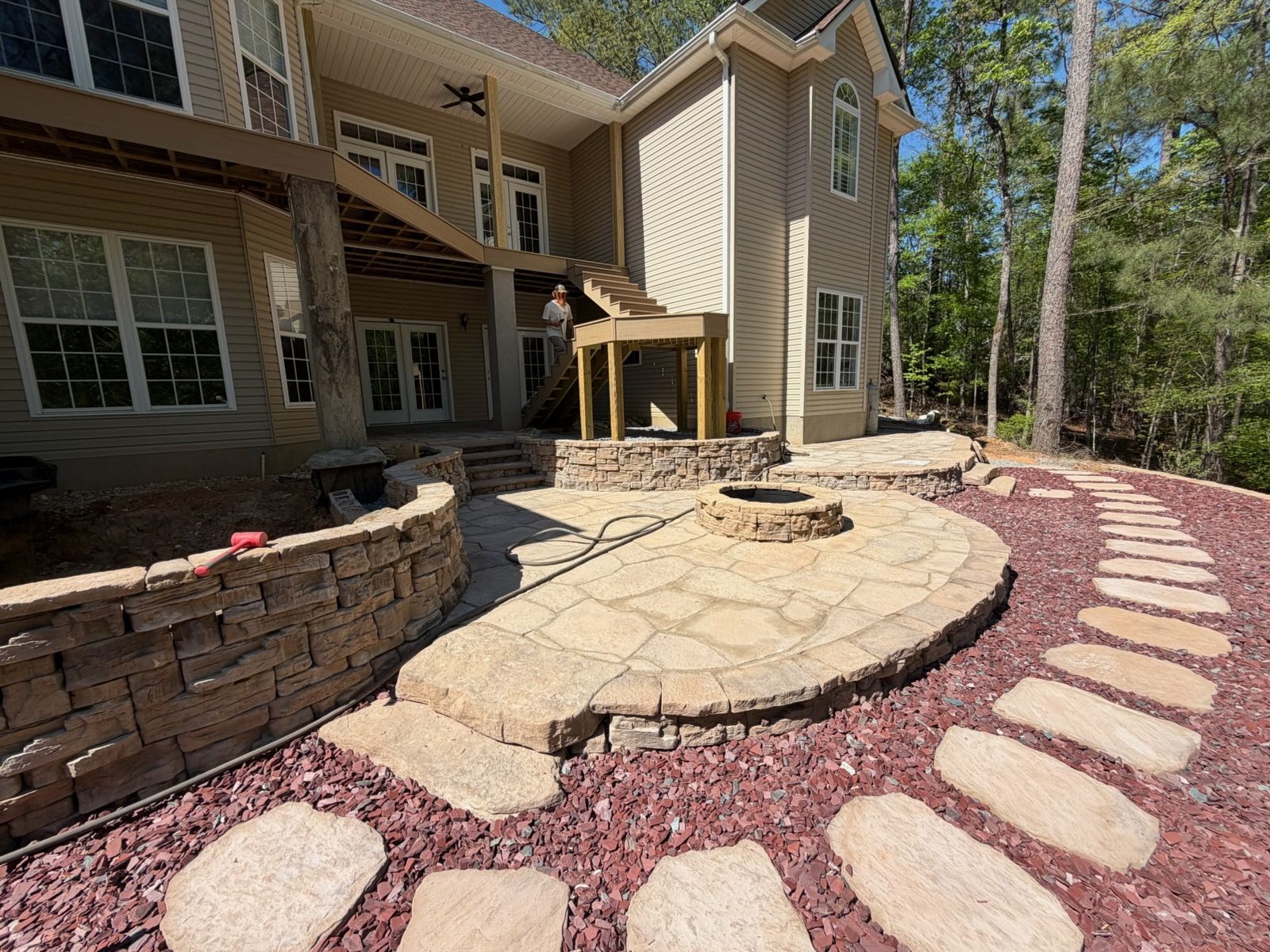 Person tending plants in a backyard pond with rocks, tall grasses, and a house in the background