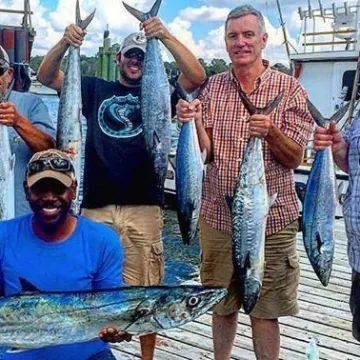 Four people hold freshly caught mackerel fish on a wooden dock with a boat in the background.