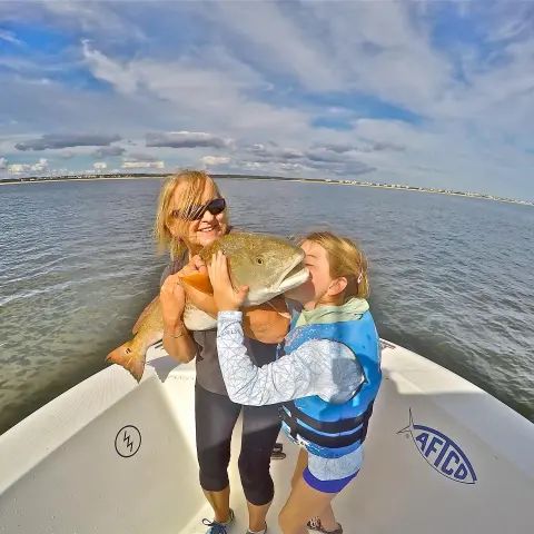 A person and a child wearing a life vest hold a large fish on a boat in open water under a cloudy sky.