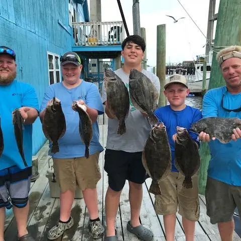 Five people standing on a wooden dock holding freshly caught flatfish in front of a blue building.