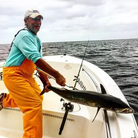 A person in a blue shirt and orange overalls holds a large fish on a boat in the open water.