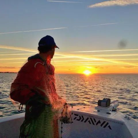 A person in a cap and red jacket stands on a boat at sunset, holding a fishing net over the water.