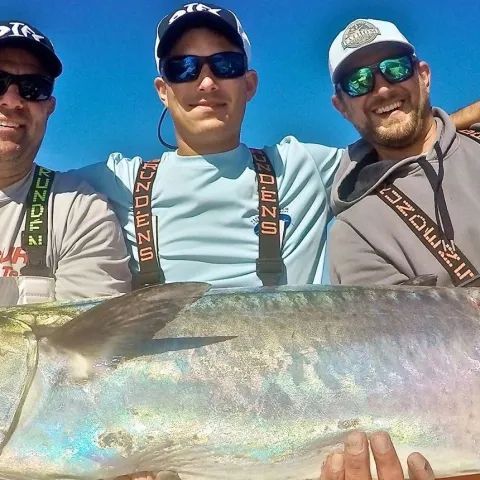 Three people wearing sunglasses and hats pose outdoors while holding a large, silvery fish.