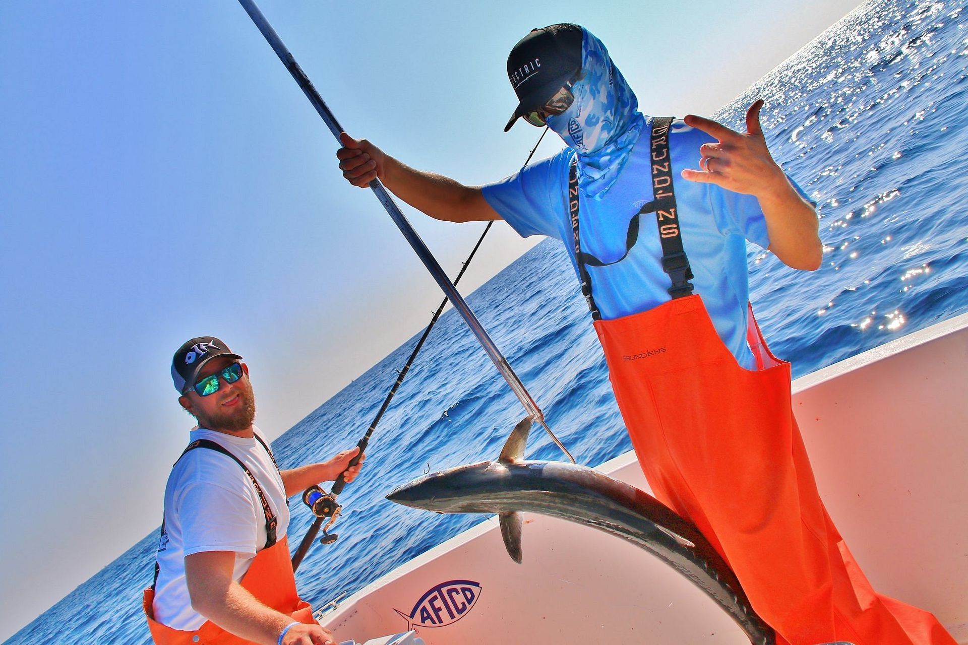 Two anglers in orange bibs and blue shirts on a boat, one holding a fishing rod and the other gesturing by a caught fish.