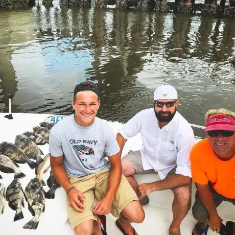 Three people pose on a boat next to a pile of freshly caught fish, with a dock and water in the background.