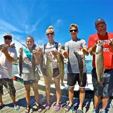 Four people stand on a wooden dock under a clear blue sky, each holding a fish they have caught.