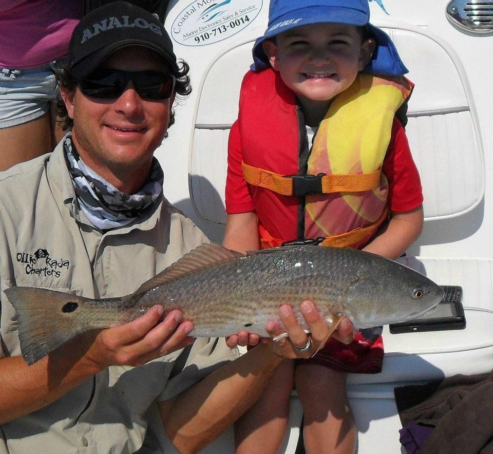 An adult and child hold a redfish while sitting on a boat, both smiling for the camera.