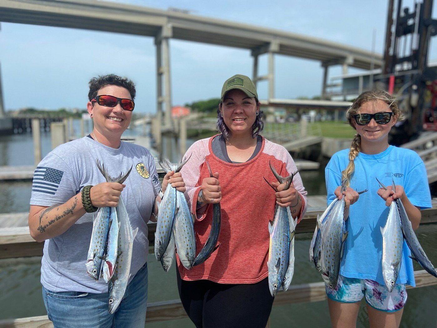Three people holding freshly caught fish on a wooden pier with a bridge and industrial equipment in the background.