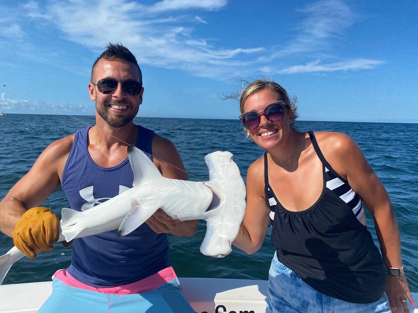 A person wearing gloves holds a small hammerhead shark next to a smiling person on a boat in the open ocean.