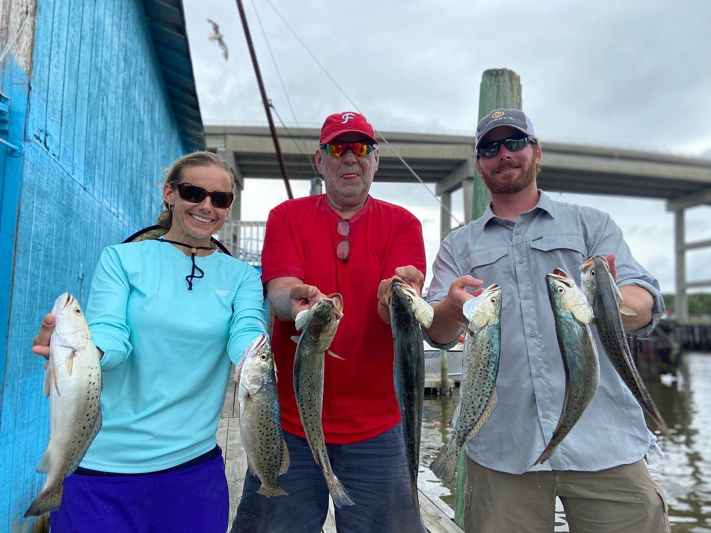 Three people stand on a dock in front of a blue building, smiling and holding up caught fish.