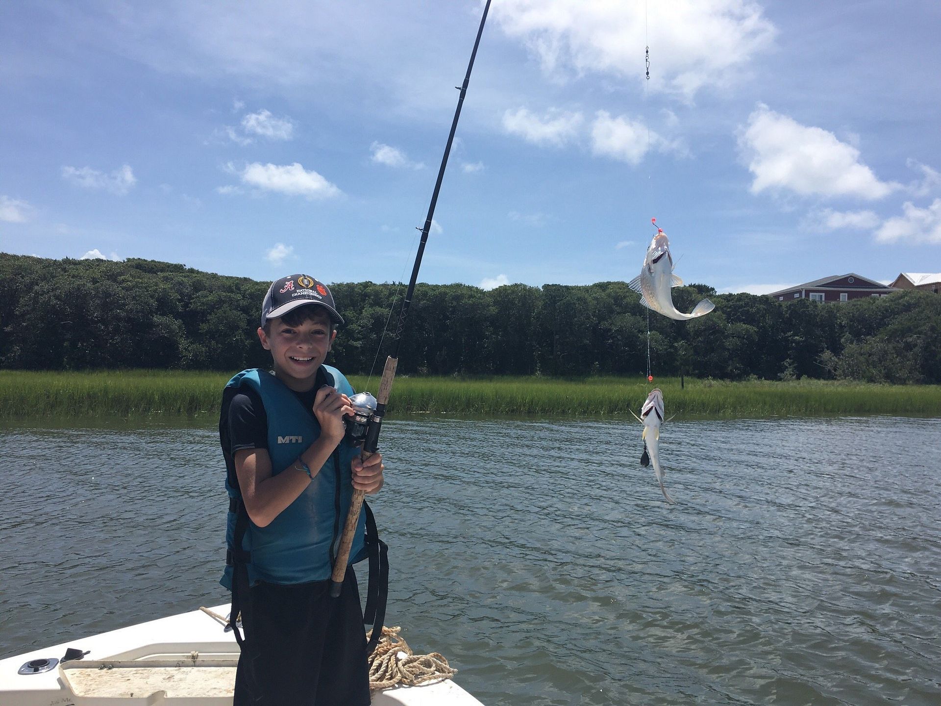 A person wearing a life vest smiles while holding a fishing rod on a boat, having caught two fish on the same line.