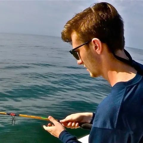 A person wearing sunglasses fishes from a boat on calm, blue open water.