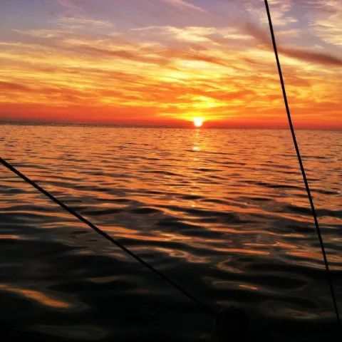A vibrant orange and gold sunset over a calm ocean, viewed from the deck of a boat with rigging visible in the foreground.