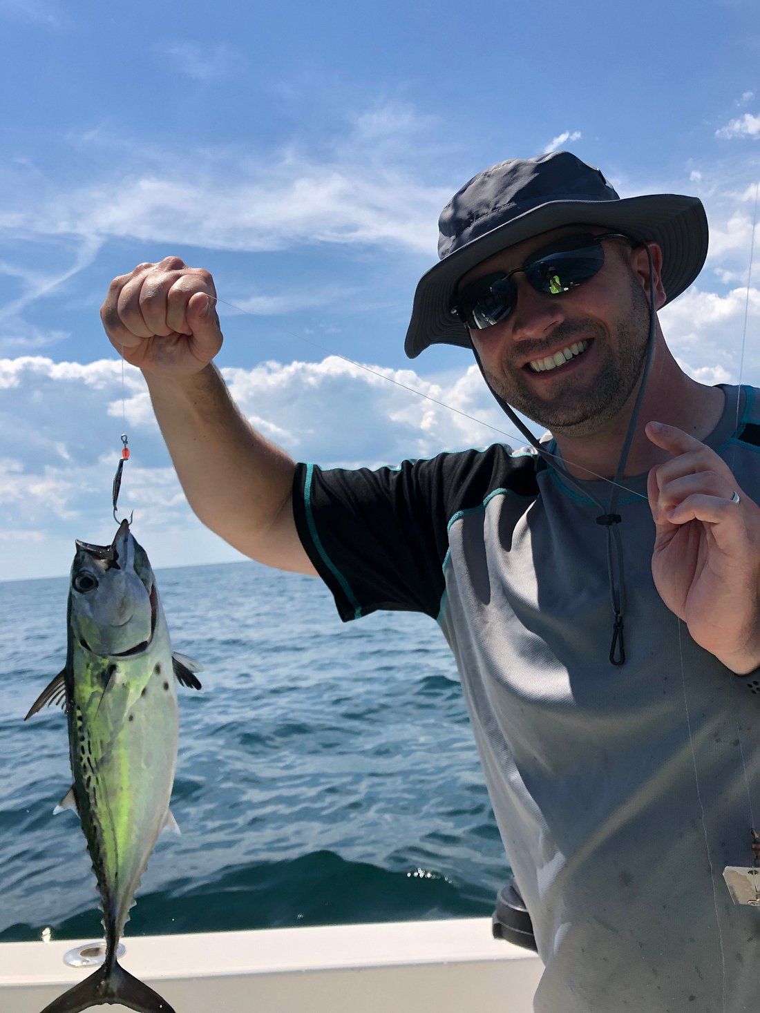 A smiling person in a hat and sunglasses holds a freshly caught fish against a sunny, blue-sky ocean background.