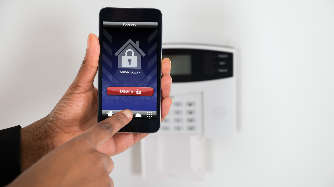 A person is holding a cell phone in front of a burglar alarm
