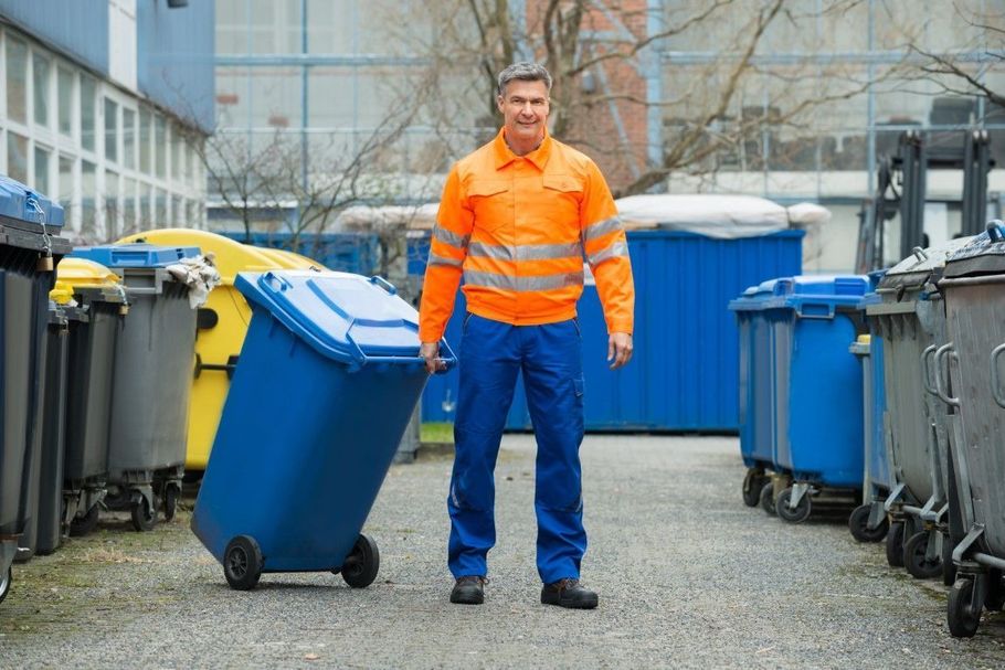 Worker in orange safety vest standing between blue recycling bins outdoors