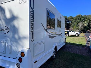 White Travel Caravan Parked On Grass Beside A Road, With Windows And A Wooded Background — Car Detailing Mobile Cleaning Co In Port Macquarie, NSW