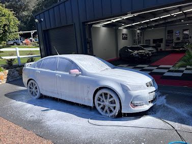 White Sedan Covered In Foam At A Car Wash Bay, With A Garage And Another Car In The Background — Car Detailing Mobile Cleaning Co In Port Macquarie, NSW