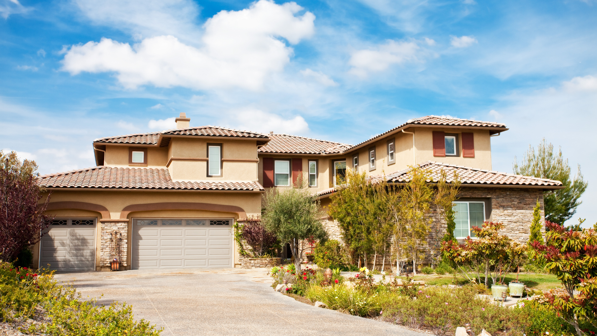 A two-story suburban home with tan stucco, a tiled roof, a multi-car garage, and landscaping under a blue sky.