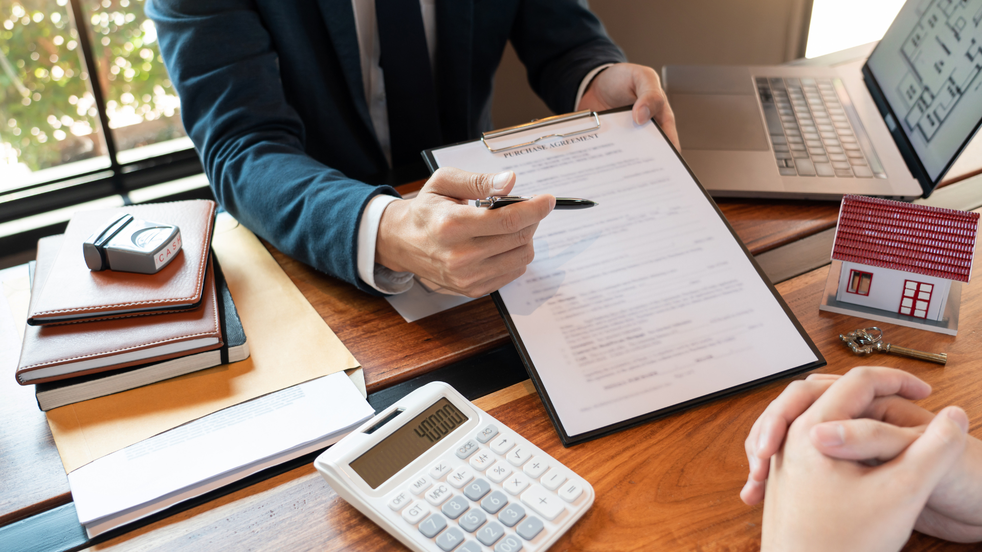A person in a suit sits at a desk with a document, calculator, and miniature house, consulting with another person.