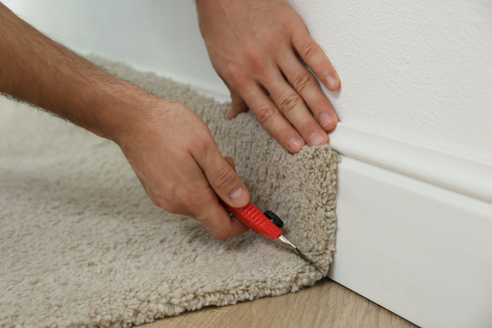 Person cuts carpet near a white baseboard with a utility knife, indoor setting.