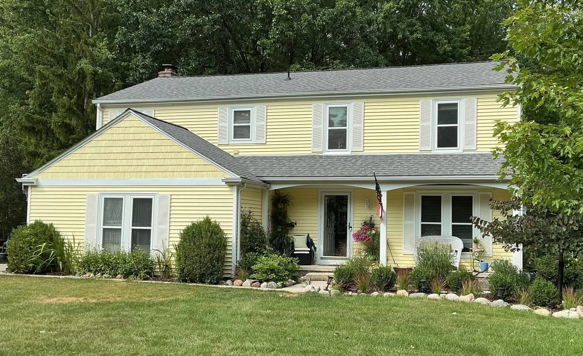 Yellow two-story house with white trim, dormer windows, and a front porch amid green trees and lawn.