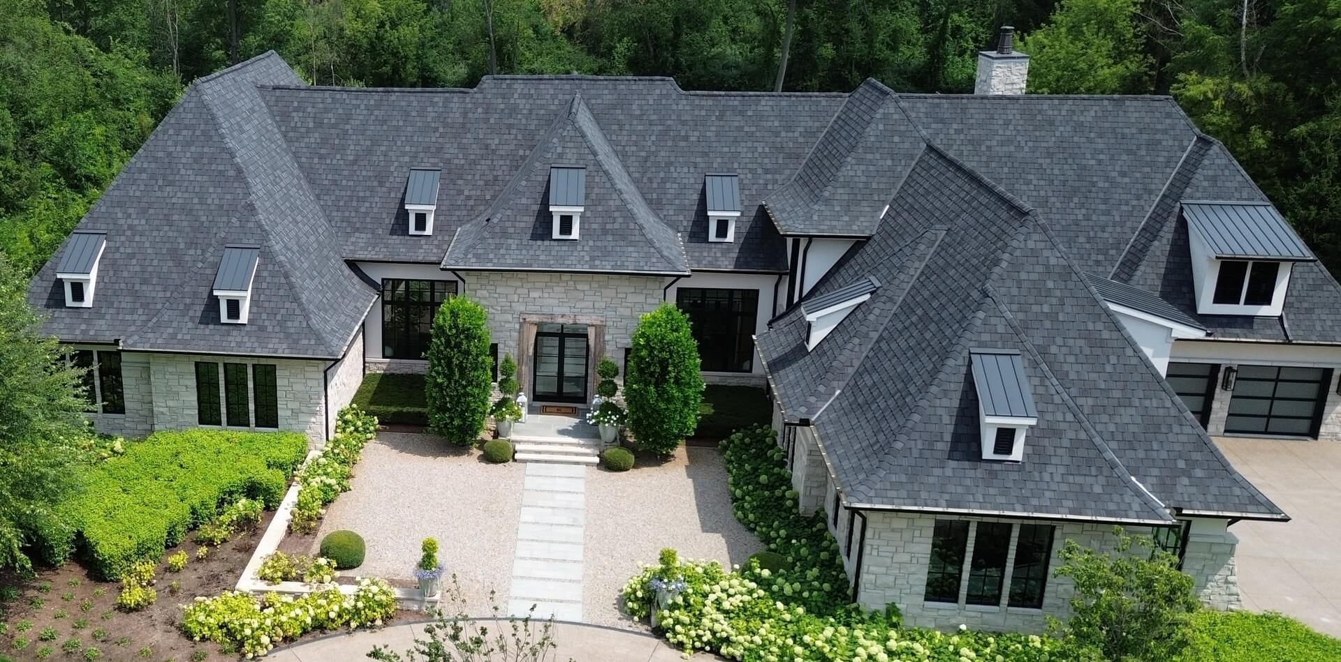 Large gray stone house with a dark roof, central walkway, and landscaped front yard viewed from above.