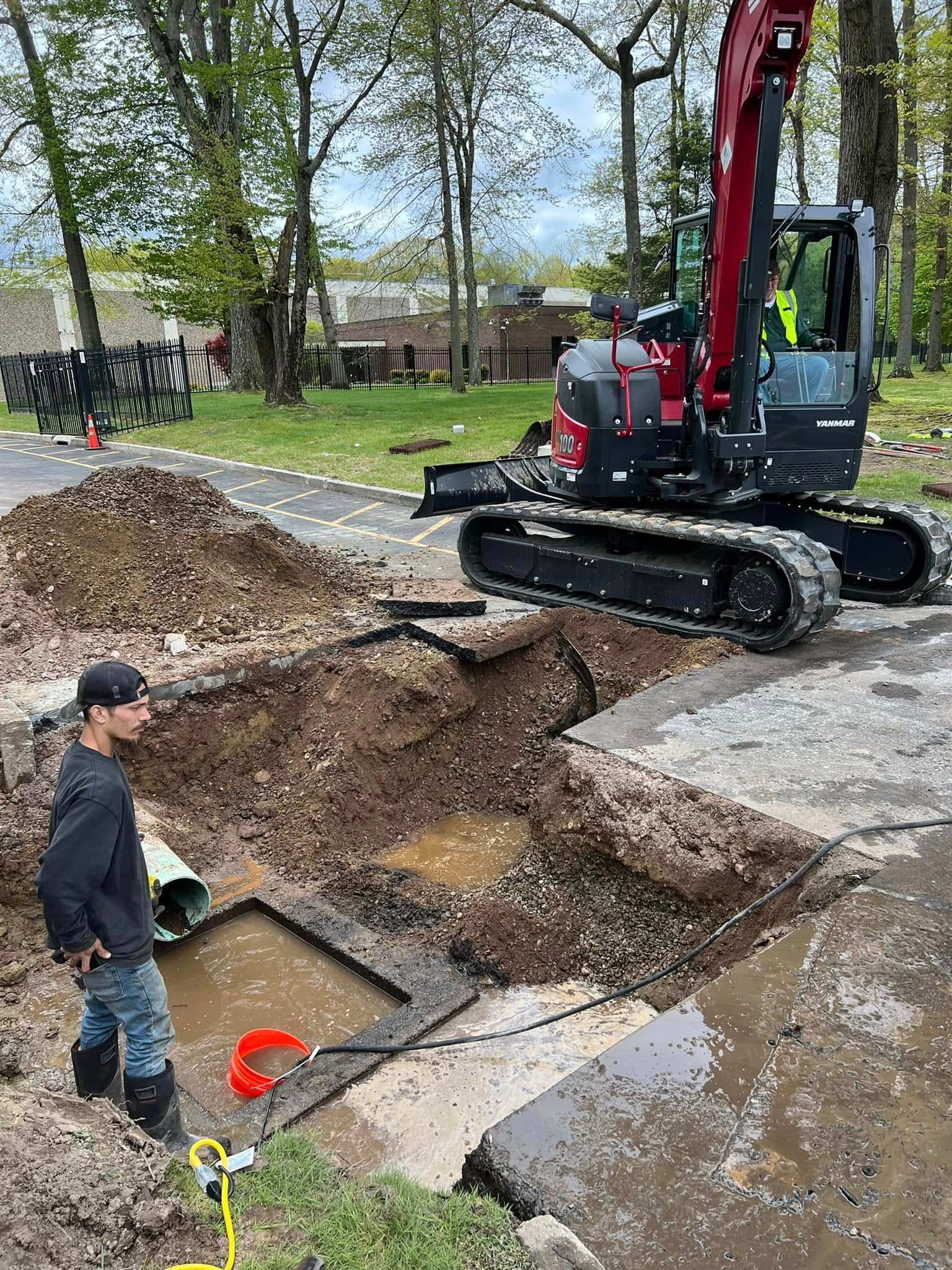 A person in a work shirt and jeans stands by a dirt trench next to a red excavator parked on a paved path. A person in a work shirt and jeans stands by a dirt trench next to a red excavator parked on a paved path.