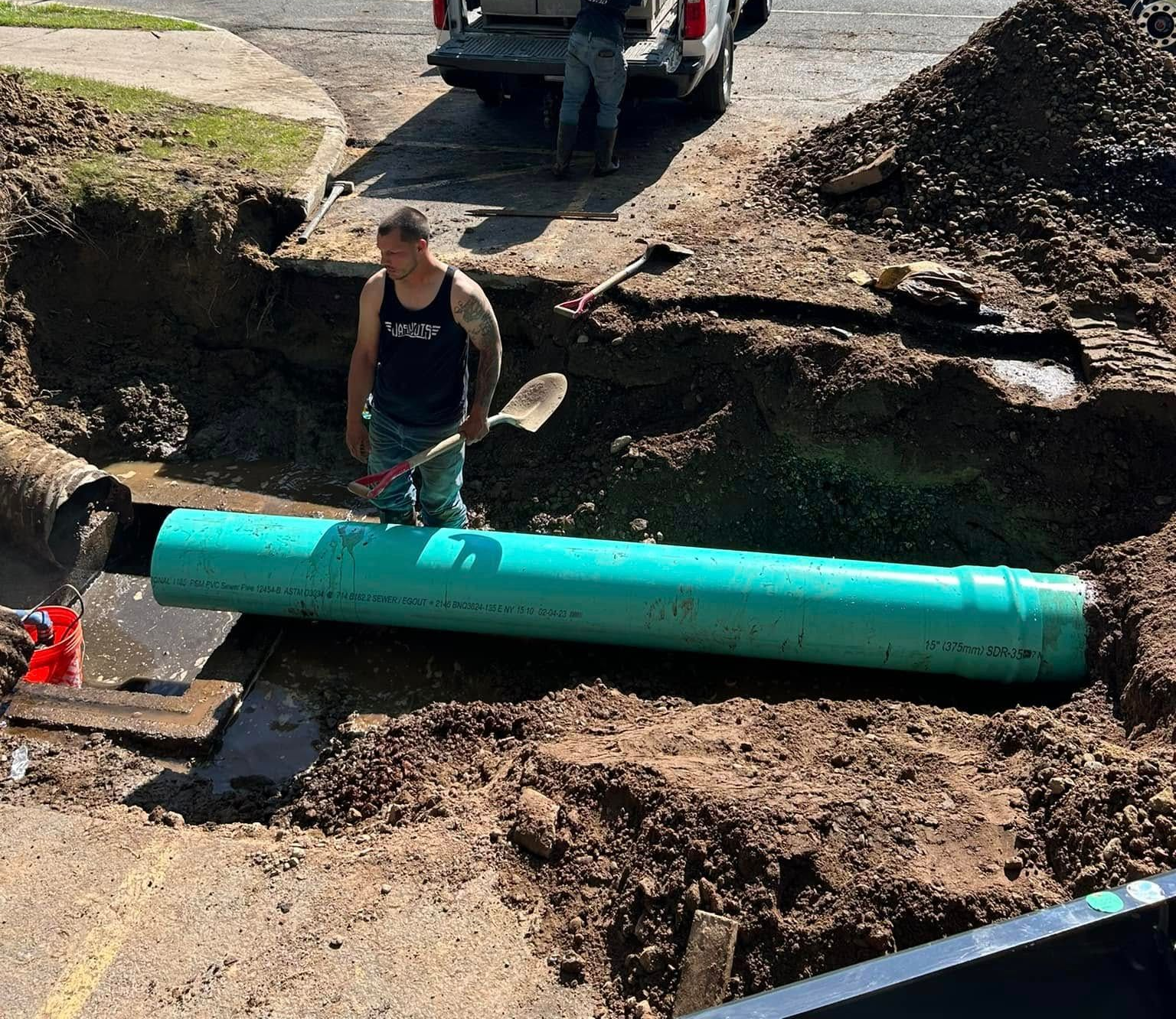 A worker stands in a trench next to a large green pipe being installed underground. A worker stands in a trench next to a large green pipe being installed underground.