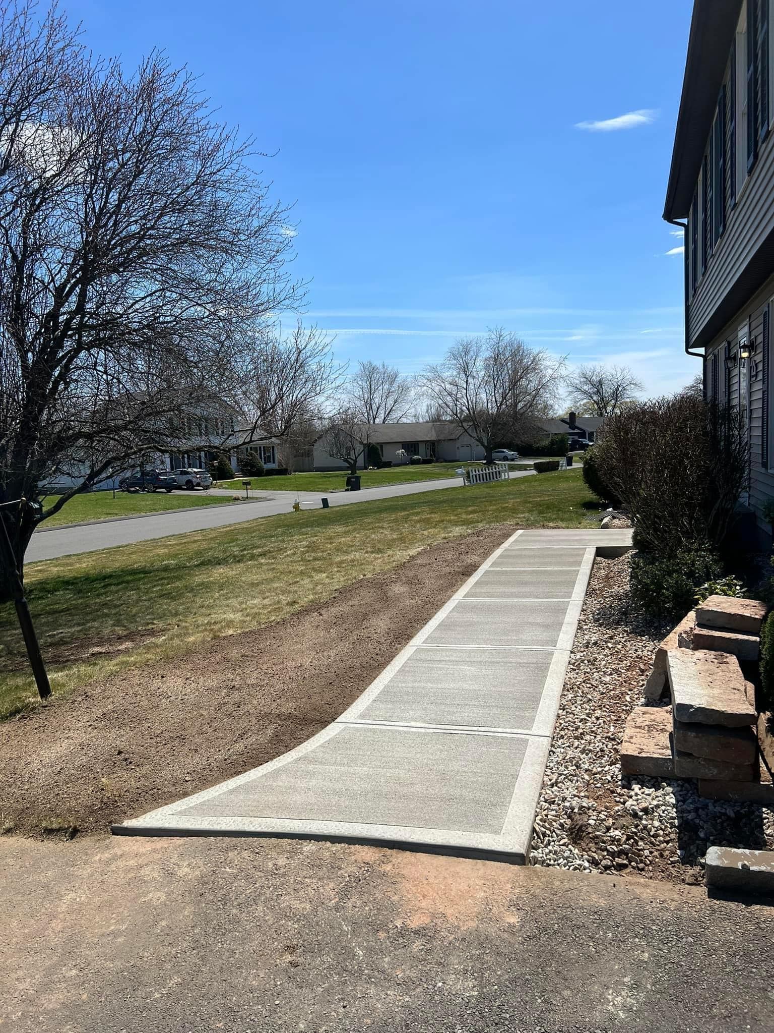 A concrete walkway leads from a driveway toward the side of a suburban house, flanked by a grassy lawn and landscaping rocks. A concrete walkway leads from a driveway toward the side of a suburban house, flanked by a grassy lawn and landscaping rocks.
