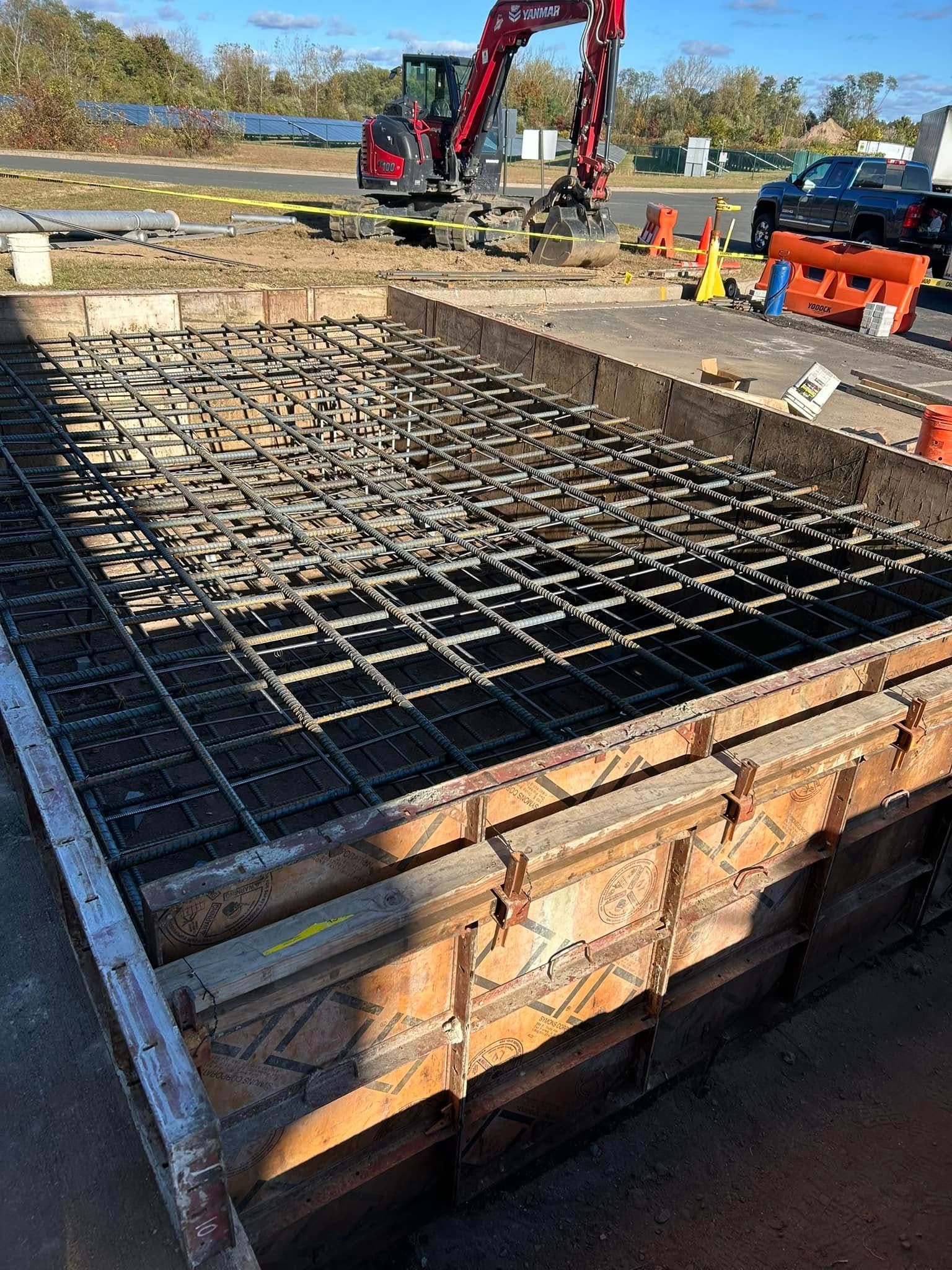 A reinforced concrete foundation form filled with a metal rebar grid at a construction site with an excavator nearby. A reinforced concrete foundation form filled with a metal rebar grid at a construction site with an excavator nearby.