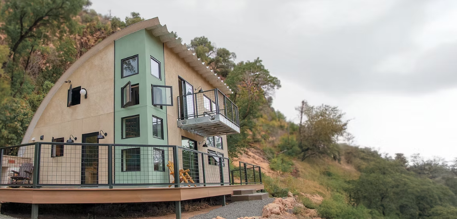 Modern hillside house with glass balconies on a wooden deck beside a rocky, tree-covered slope