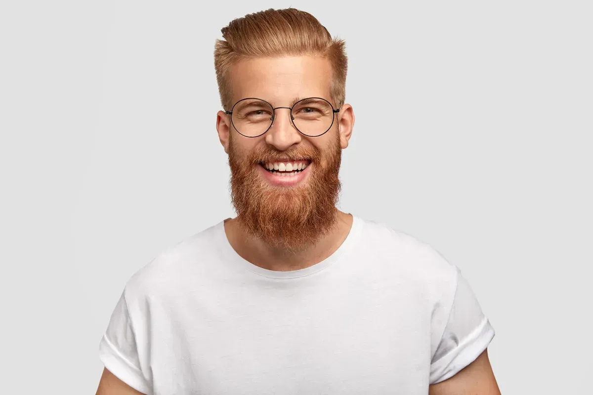 Man with red beard, glasses, and white shirt smiling against a light gray background.
