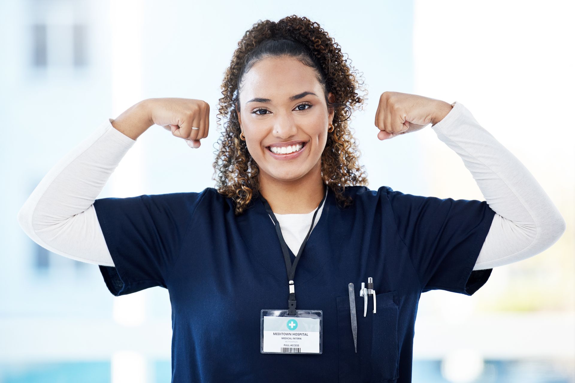 Smiling person in scrubs flexing, with badge and pens, lit room.