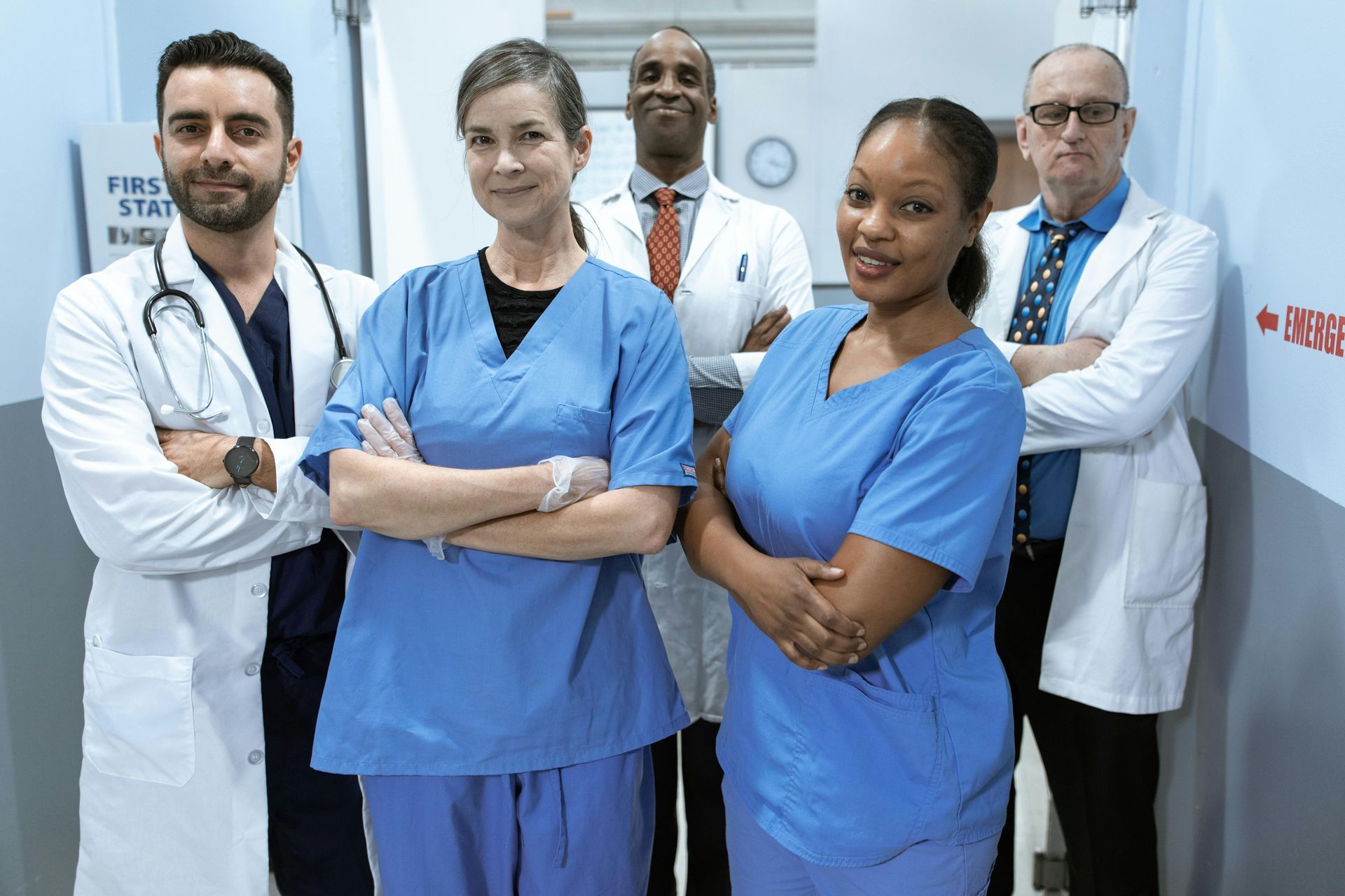 Four hospital staff in blue scrubs and white coats standing in a clinic hallway, arms crossed