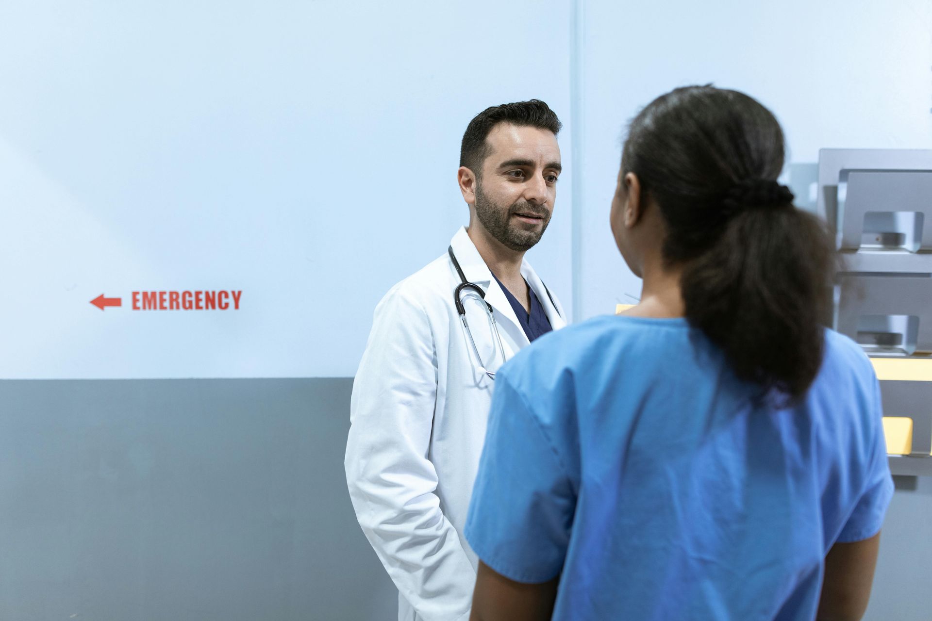 Doctor in white coat speaking to a person in blue scrubs near an emergency room door.