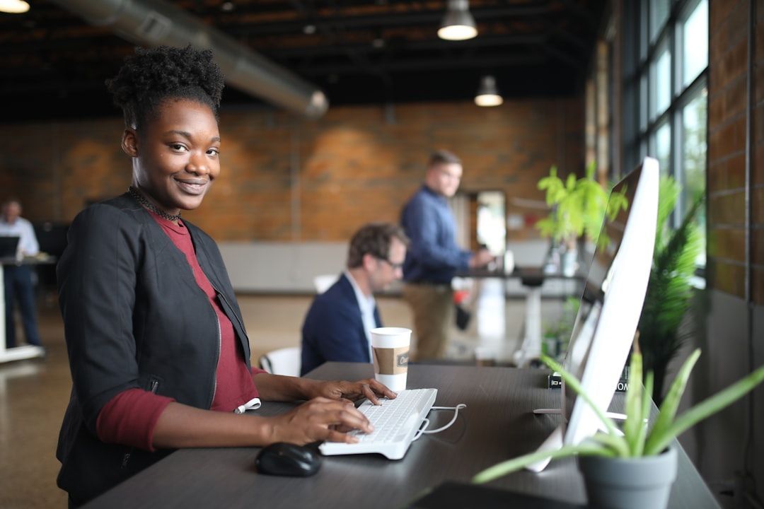 Person smiling at a desk with a laptop in a bright office, with coworkers blurred in the background