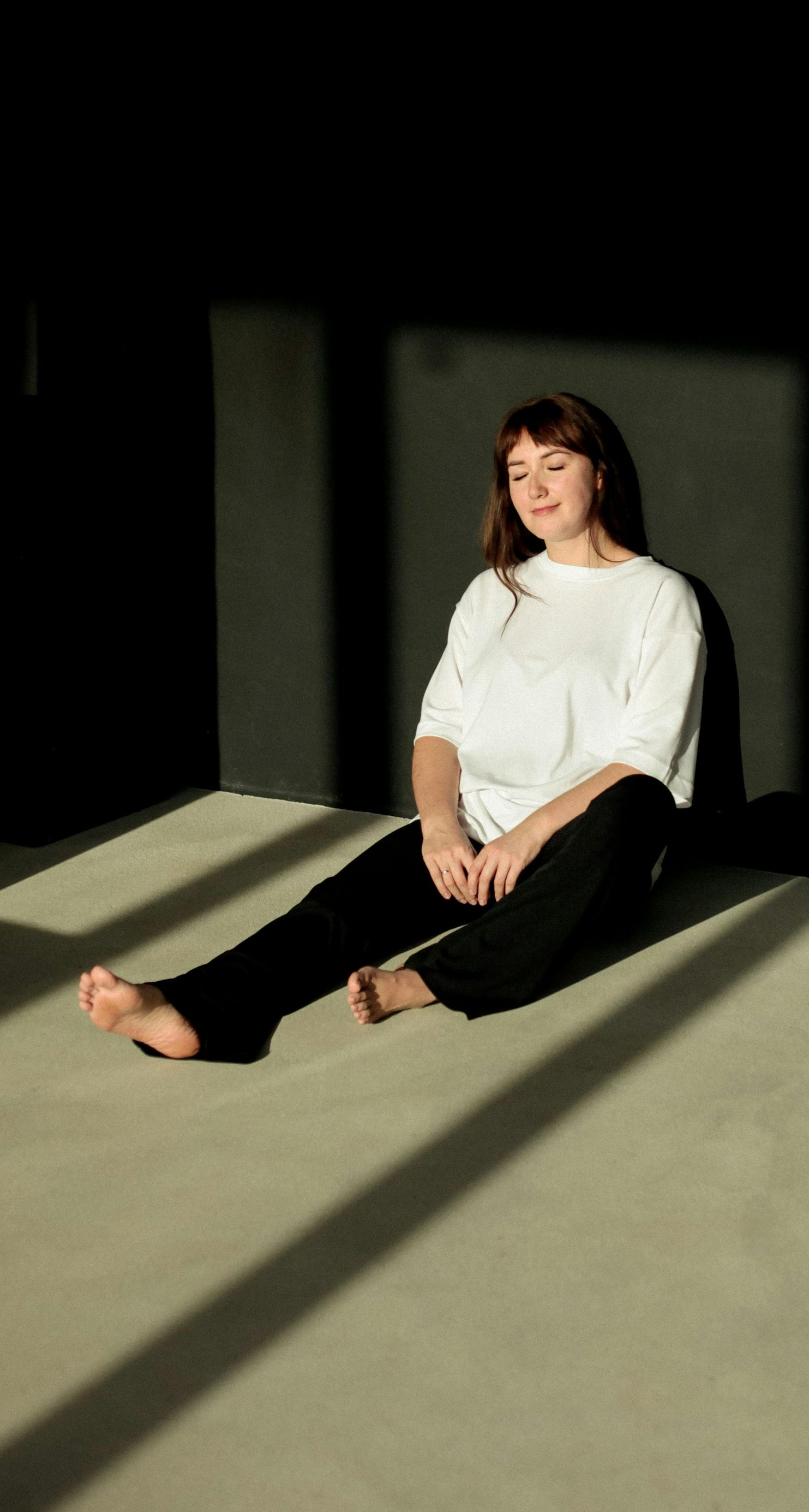 Woman sits on floor in sunlight, leaning against a dark wall, wearing white shirt and black pants.