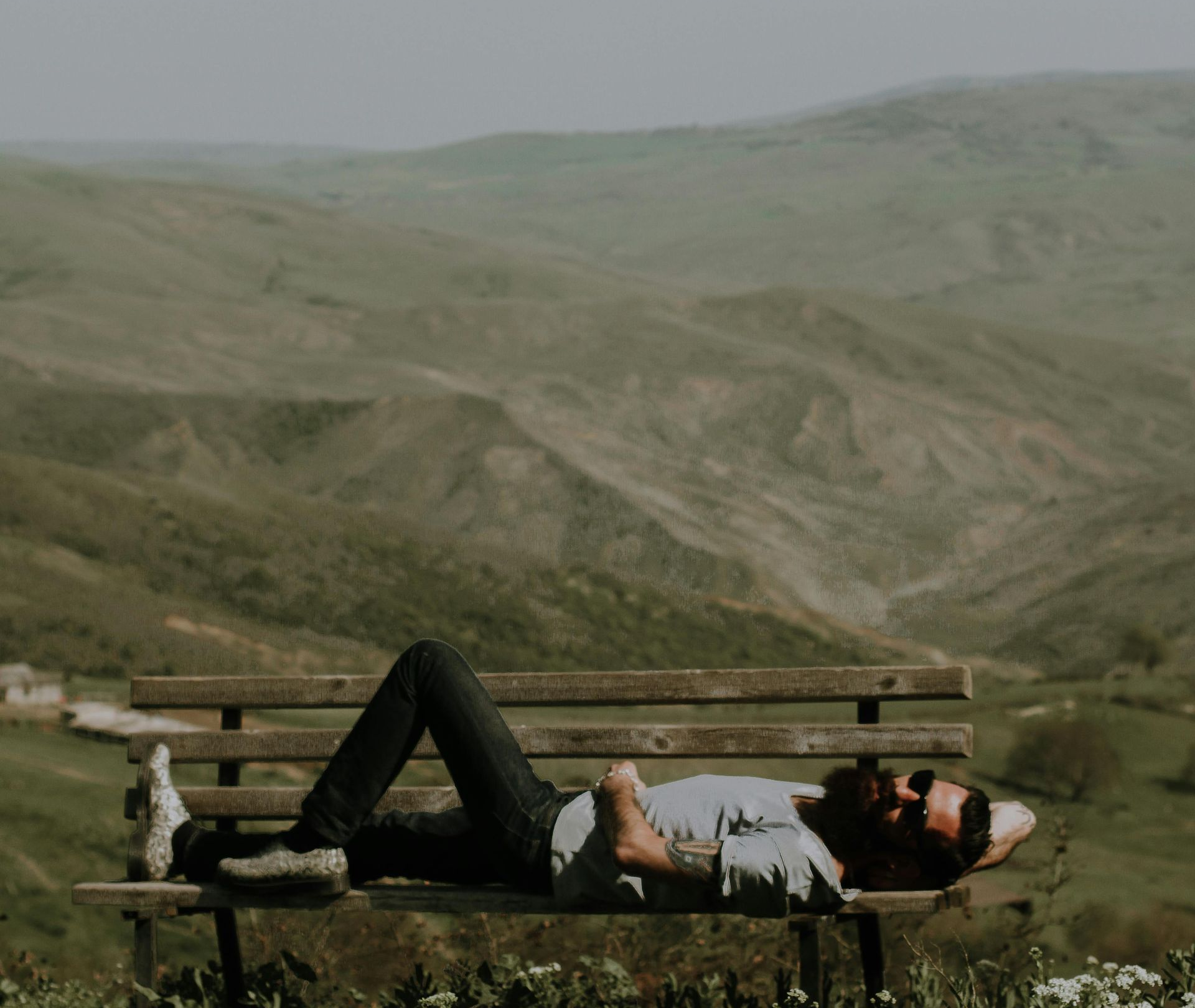Man with beard reclines on a wooden bench, overlooking a hilly, green landscape.